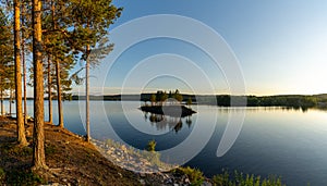 Panorama of a calm lake with small island and golden sunset evening light on the trees and forest on the lakeshore in the