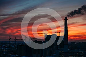 Panorama of city sunset and silhouettes of cranes, high-rise buildings and construction site with smoke