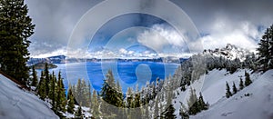 Panorama of Crater Lake with deep blue water, grand cloudy sky and snow covered trees in the foreground