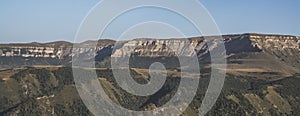 Panorama in the evening in the mountains of the Caucasus on a steep rocky slope of a table mountain and wooded hills with trees