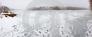 Panorama of a frozen river foggy winter day. Fisherman on ice in