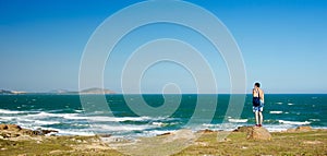 Panorama. A man with shorts with a bag on his shoulder is standing on a rock and looking at the sea