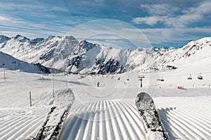 Panorama point of view skier legs on downhill start straight line rows freshly prepared groomed ski slope piste on bright day blue