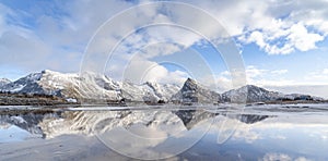 Panorama reflection of snowy mountains, blue sky and clouds on the calm clear water on a beach.