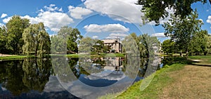 Panorama of Stewart Park, a view of the willow trees and stone house, surrounded by other trees, reflected on pond in