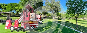 Panorama view firetruck themed playground with green artificial grass turf carpet by tall mature trees clear blue sky in