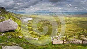 Panorama with view on steep stairs of wooden boardwalk leading to Cuilcagh Mountain peak with lake and valley below