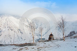 Panoramic aerial top view to winter Caucasian mountains and glaciers, small trees and a grave near Gergeti Trinity church