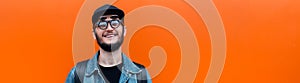 Panoramic banner, portrait of smiling millennial hipster guy wearing denim jacket, cap, backpack and glasses orange background.