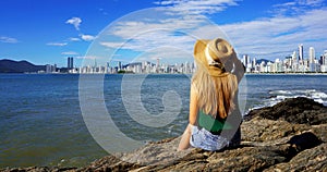 Panoramic banner view of young woman sitting on the rocks looking at Balneario Camboriu skyline on Atlantic Ocean. Summer vacation