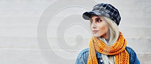 Panoramic outdoor profile portrait of beautiful young blonde girl with blue eyes wearing orange scarf, denim jacket and plaid cap.