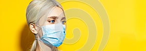 Panoramic portrait of young blonde girl with blue eyes, wearing medical face mask against coronavirus on yellow background.