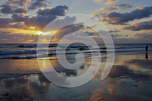 Panoramic scene of ocean sunset with clouds reflected on wet beach and person wading