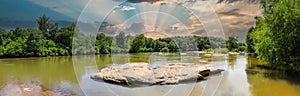 A panoramic shot of the Chattahoochee river with rocks on the middle of the river and green trees reflecting off the water