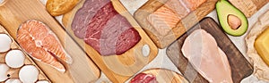 Panoramic shot raw meat, fish and poultry on wooden cutting boards near chicken eggs and half of avocado.