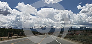 Panoramic shot of the road between the fir forest trees and mountains against a blue clouded sky