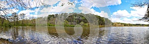 A panoramic shot of a vast rippling lake surrounded by lush green trees with powerful clouds and blue sky at Murphey Can