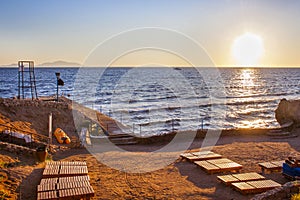Panoramic view of an empty sandy beach with sunbeds and sunset over the sea. The sun sets behind the horizon, forming a path on th