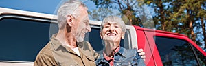 panoramic view of happy senior couple of tourists standing near car and embracing with smile.