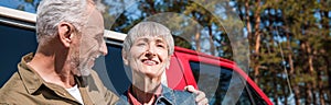 panoramic view of happy senior couple of tourists standing near car and embracing with smile.