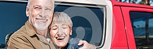 panoramic view of happy senior couple of tourists standing near car and embracing with smile.