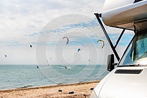 Panoramic view of many surf board kite riders on sand beach watersport spot on bright sunny day against rv camper van