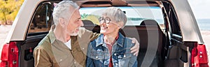 panoramic view of smiling senior couple embracing and looking at each other near car.