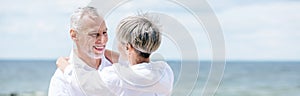 panoramic view of smiling senior couple embracing near river under blue sky.