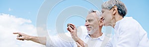 panoramic view of smiling senior couple in white shirts looking away and pointing with finger under blue sky.