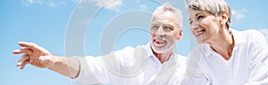 panoramic view of smiling senior couple in white shirts looking away under blue sky.