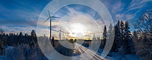 Panoramic wide scenic aerial photo over forest winter road with windmills standing in row at the left side in forest. Road at the