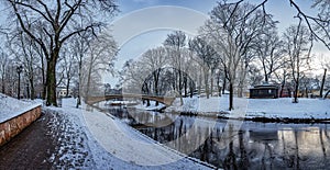 Panoramic winter landscape in snowy park with beautiful bridge over small canal, street light and covered in snow tress. View of