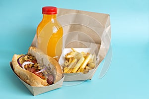 A paper bag, a fresh hot dog with sausage, French fries and a bottle with an orange drink on a blue