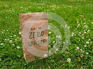 A paper bag with handwritten words `Go to zero waste` on it among clover and green grass with copy space.
