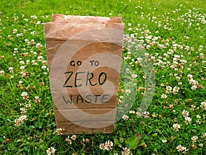A paper bag with handwritten words `Go to zero waste` on it stands among clover and green grass.