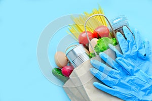 Paper bag with various food on a blue background: vegetables, eggs, buckwheat, canned goods, pasta, lettuce and rubber gloves. Top