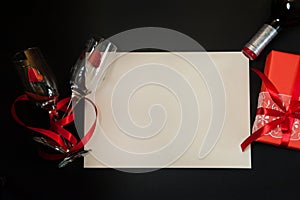 Paper blank, two narrow empty glasses intertwined with a red ribbon, a gift box, red hearts and a bottle of wine on a black backgr