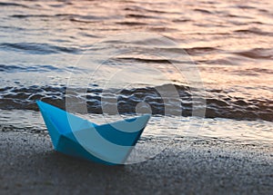 Paper blue ship is thrown on sandy shore of lake at sunset.