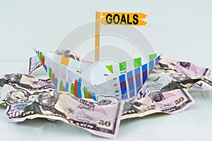 A paper boat from a business graph floats on a sea of money. The text is written on the flag - GOALS