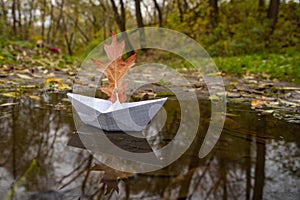 The paper boat floats on an autumn puddle, being reflected in water. Instead of a sail, a red oak leaf