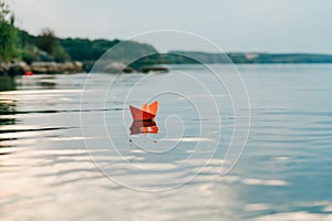 A paper boat sails by the river in the summer. It has an orange color and floats downstream along the shore