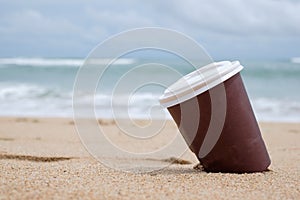 Paper brown cup of coffee on sand beach over cloudy sky and sea.