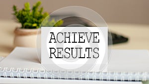Paper card with text ACHIEVE RESULTS. Calculator, flowers and brown background