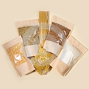 Paper-cellophane bags with cereals, legumes and pasta laid out on beige backdrop. Top view