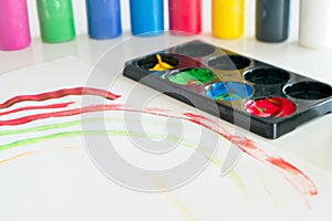 Paper with a child`s drawing with several containers of tempera paint and a plastic cup with water