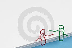 Paper clips red and green sitting on a white paper ream and talking or having a conversation. A photo with a copy space. Good for