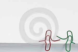 Paper clips red and green sitting on a white paper ream and talking or having a conversation. A photo with a copy space. Good for