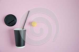 A paper coffee cup lies on a pink background. A black cover is inserted into the glass. A yellow macarone lies next to it. Blurred