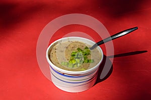 Paper container filled with brown speckled mushroom soup with chopped chives and a black plastic spoon on top isolated on a red