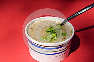 Paper container filled with brown speckled mushroom soup with chopped chives and a black plastic spoon on top isolated on a red